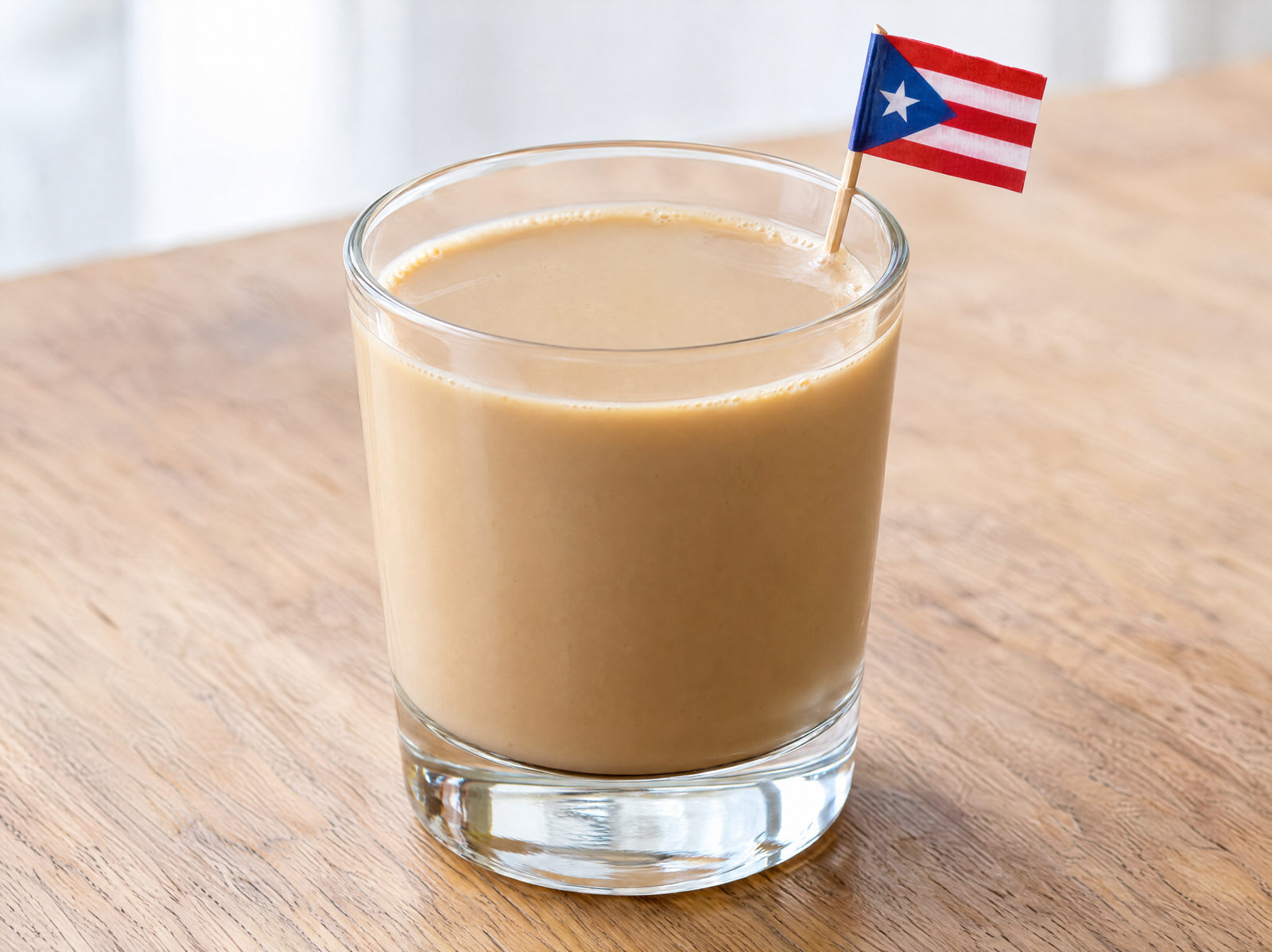 Creamy light-tan coquito de café in a small clear glass on a natural wooden table